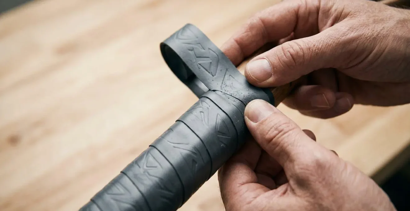 Vue rapprochée des mains d'un cordeur appliquant un surgrip sur un manche de raquette avec chevauchement précis
